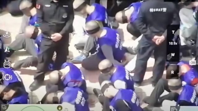 Des prisonniers Ouighours rasés, entravés et les yeux bandés, sous haute surveillance policière (capture d'écran de la vidéo Youtube "War on Fear")